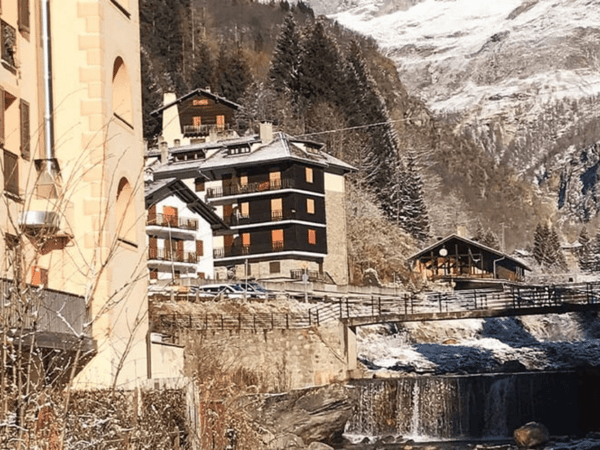 Snow-dusted alpine village with chalet-style hotel by a river and wooden bridge.