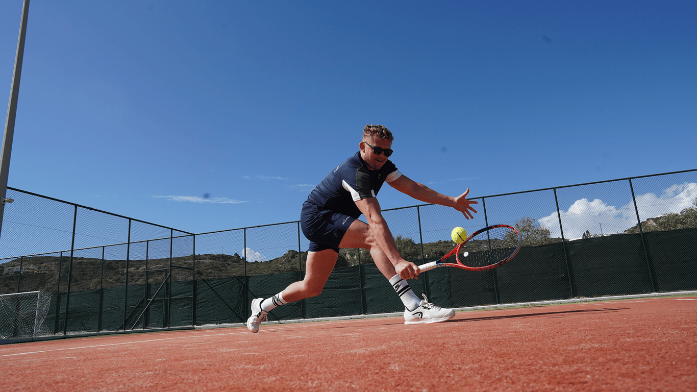 Tennis player lunges for a low forehand on a sunny clay court.