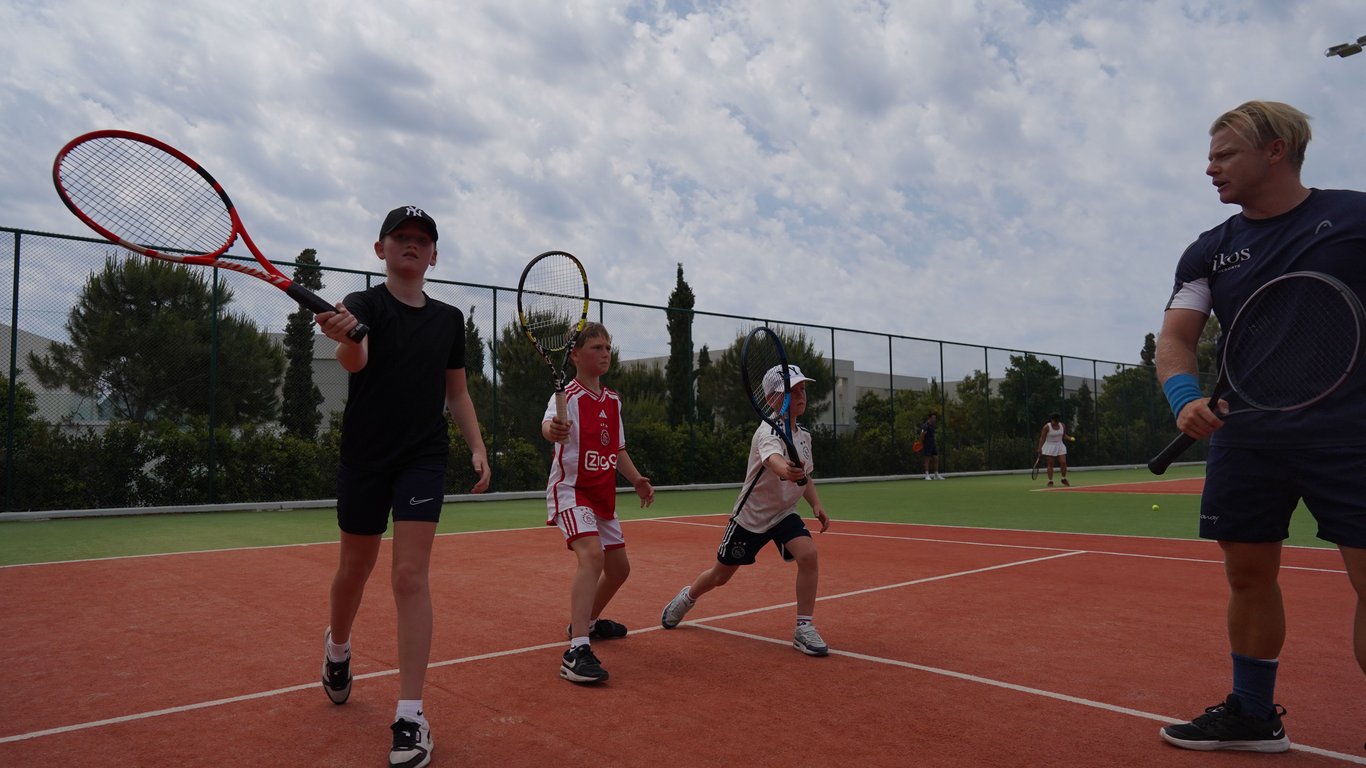 Coach guiding three children practising forehand swings on an outdoor tennis court during a lesson.
