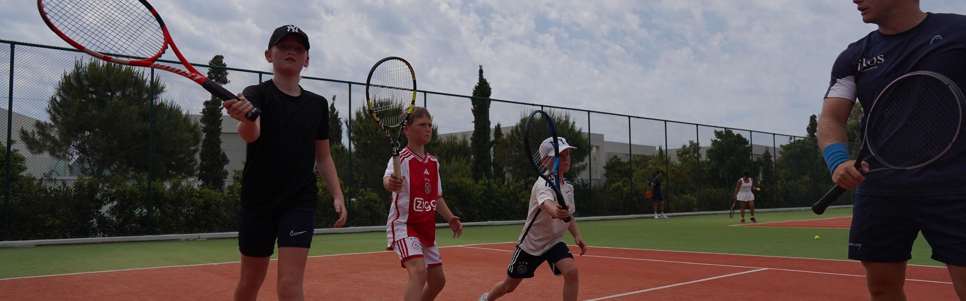 Coach guiding three children practising forehand swings on an outdoor tennis court during a lesson.