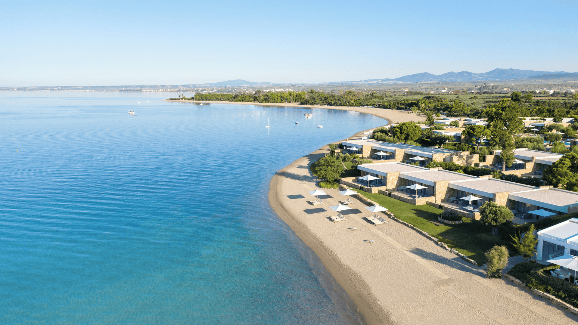 Aerial view of Ikos Resorts: beachfront villas, sandy bay, calm blue sea and sun loungers.