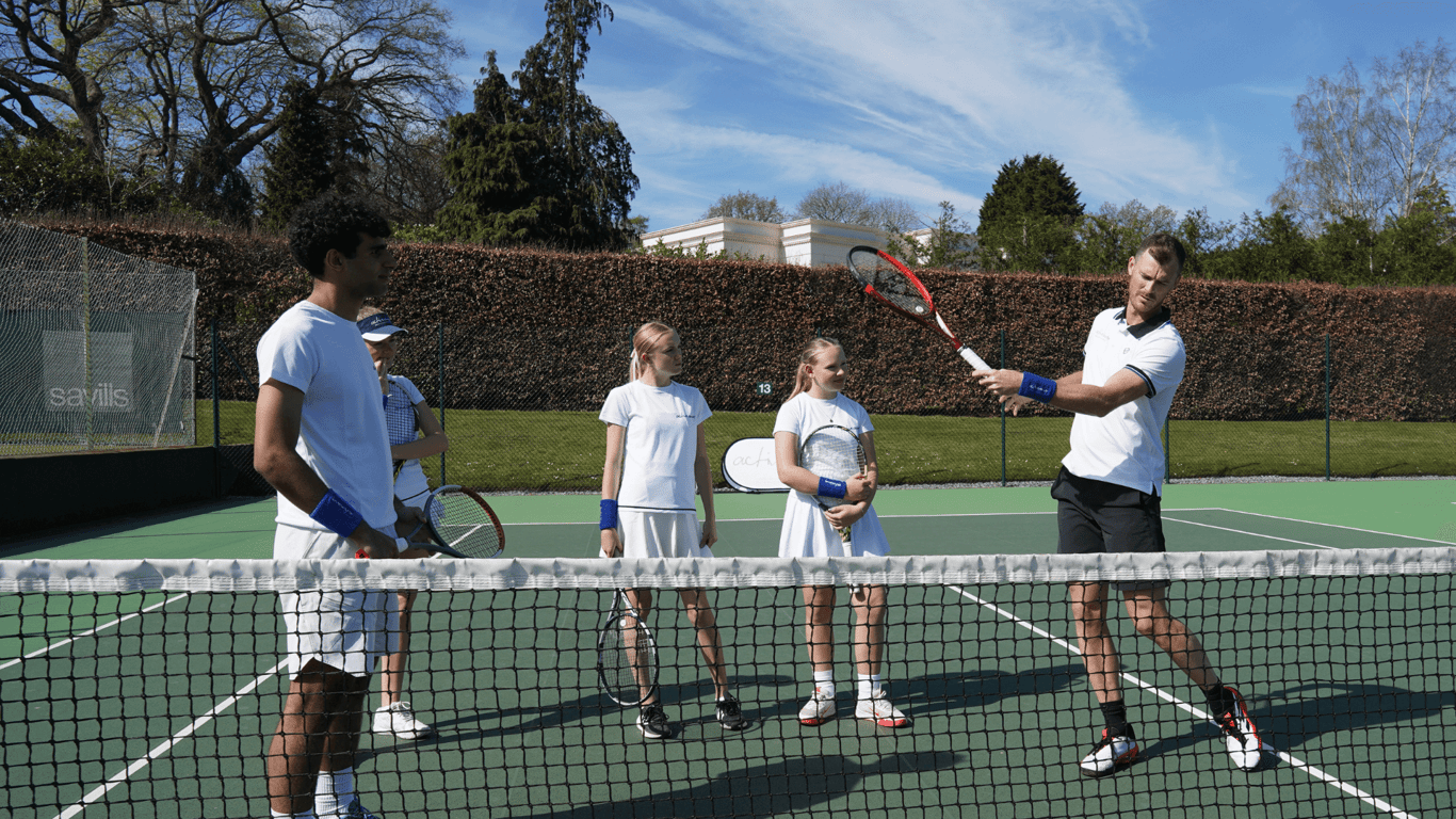 Coach demonstrates forehand to junior players during a school tennis clinic on an outdoor court.
