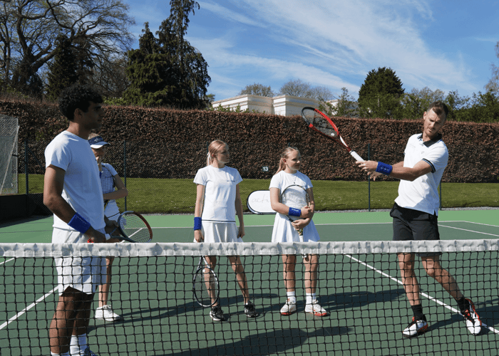 Coach demonstrates forehand to junior players during a school tennis clinic on an outdoor court.