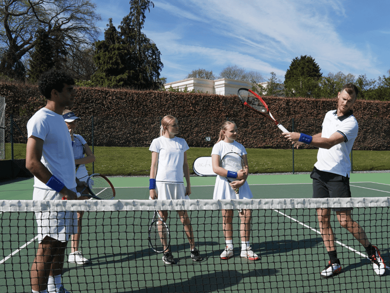 Coach demonstrates forehand to junior players during a school tennis clinic on an outdoor court.
