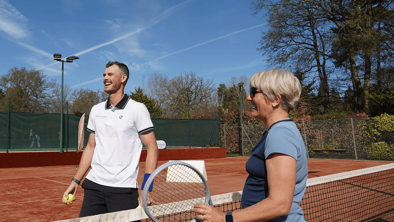 Two adults laugh by the net on a clay tennis court, holding a racket and ball on a sunny day