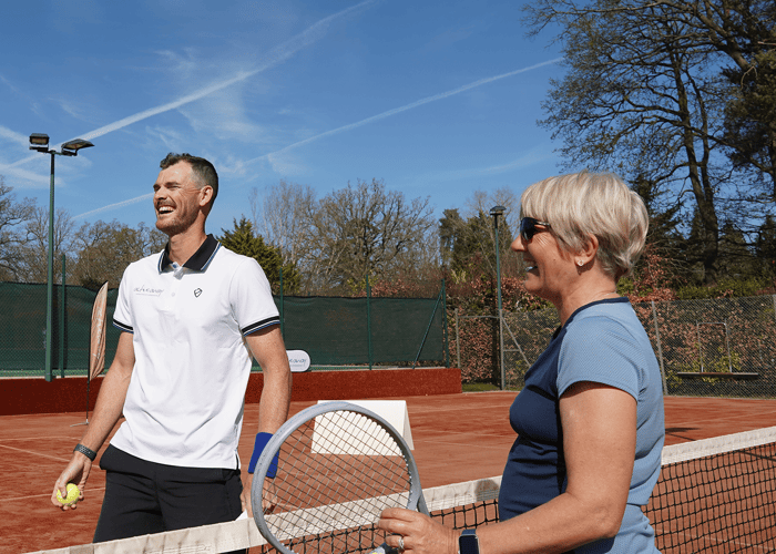 Two adults laugh by the net on a clay tennis court, holding a racket and ball on a sunny day
