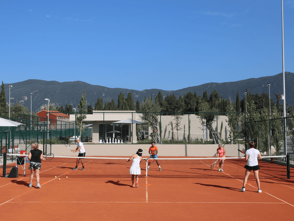 Group tennis coaching on a clay court with several players and a coach, mountains and blue sky behind.