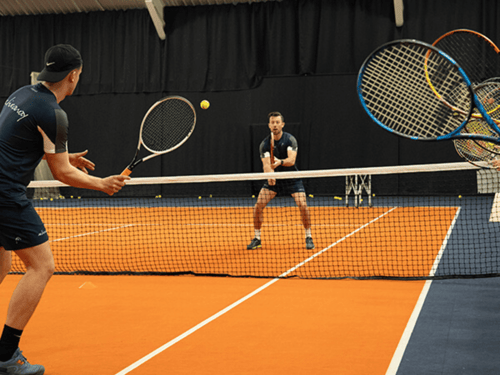 Two players practise quick volleys at the net on an indoor tennis court.