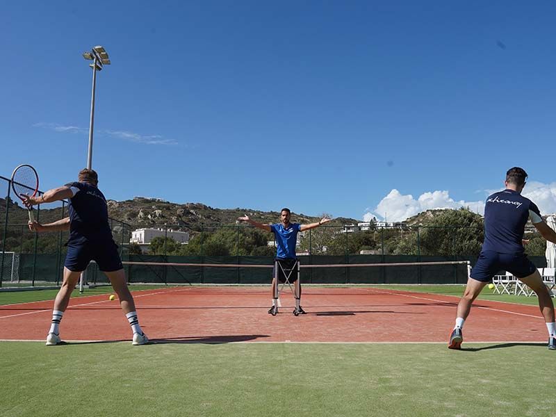 Coach feeds balls to two players on an outdoor tennis court at a sunny resort.